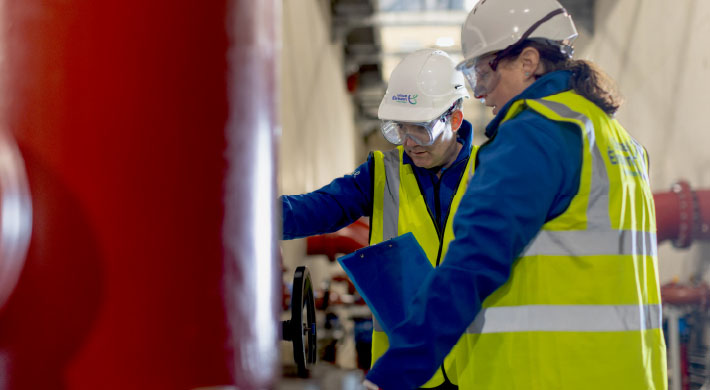 2 Wastewater treatment plant operators, male & female, discussing water flow by red intake pipes