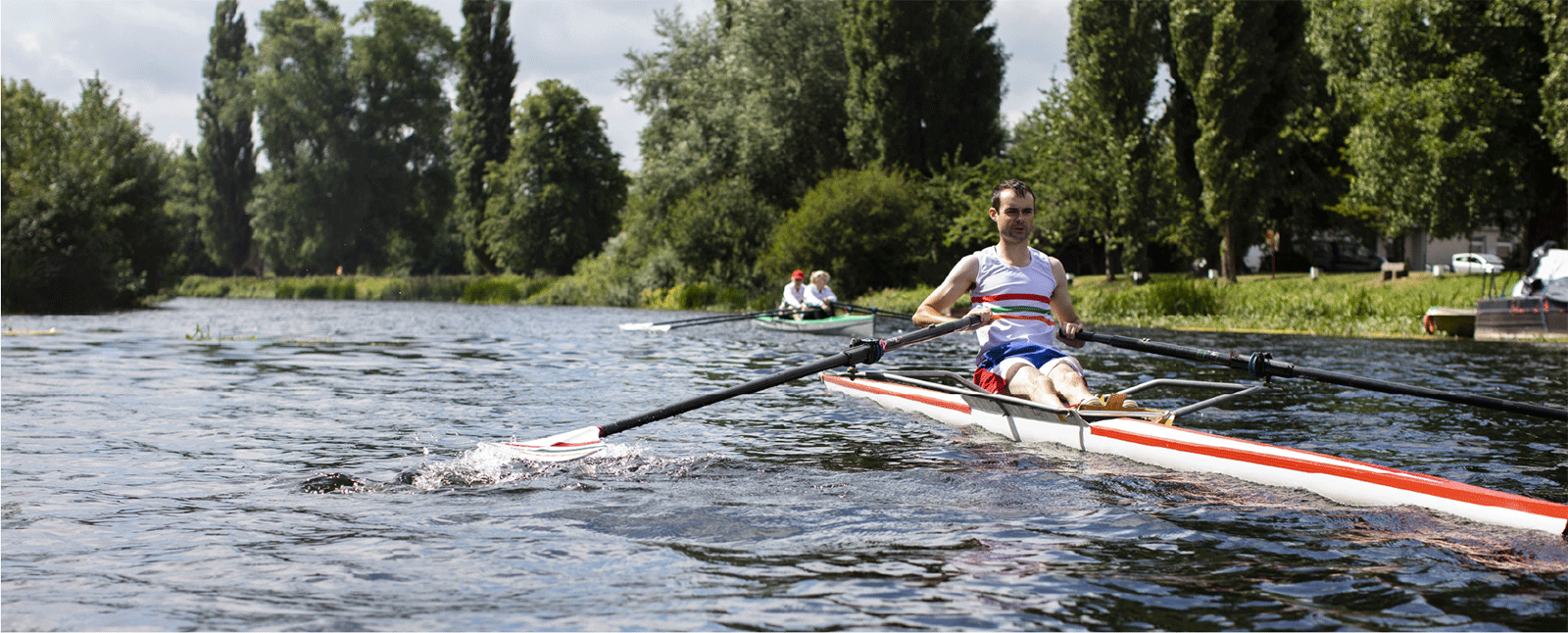 A person rowing on a calm river with trees and other rowers in the background.
