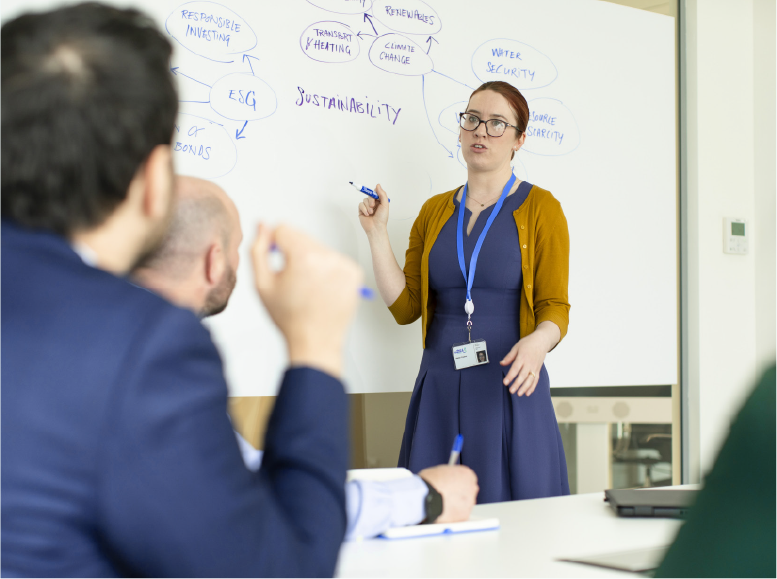 Working group environment with female standing at white board holding a marker while interactiving with seated participants.