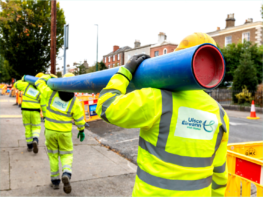 Uisece engineers in full PPE carrying a long length of piping at shoulder height at a roadside worksite.