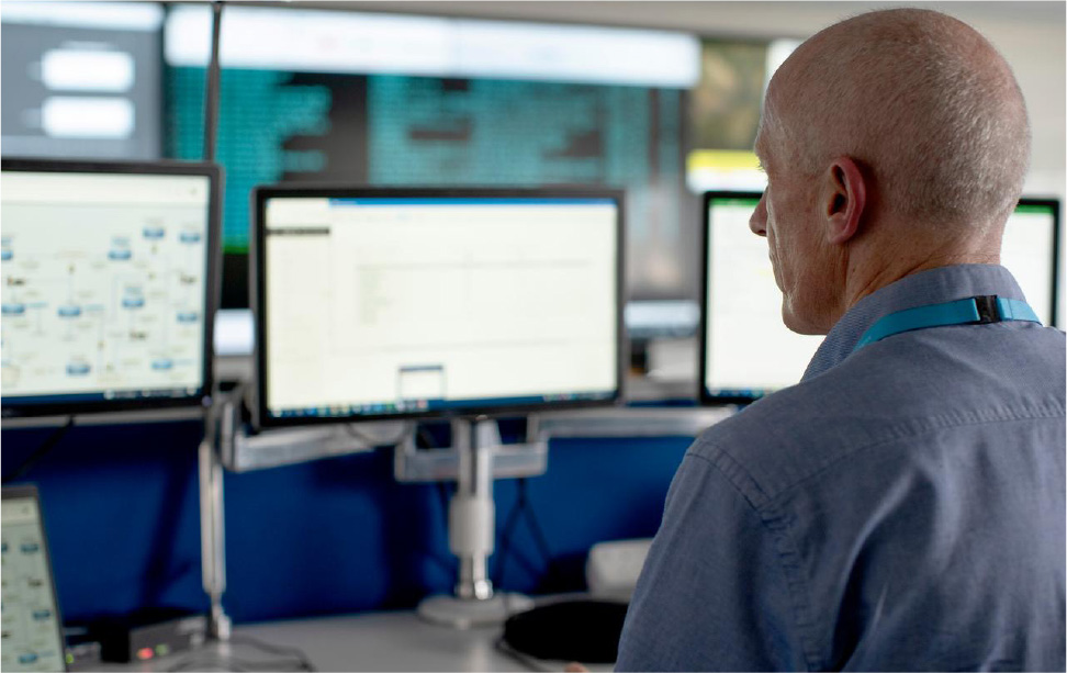 Oibrí teicniúil i suíomh monatóireachta oifige le go leor scáileán ina radharc agus scáileán balla mór sa chúlra.