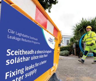 Uisce Éireann carrying a rolled up plastic blue pipe walking across an Uisce Éireann signage about fixing leaks